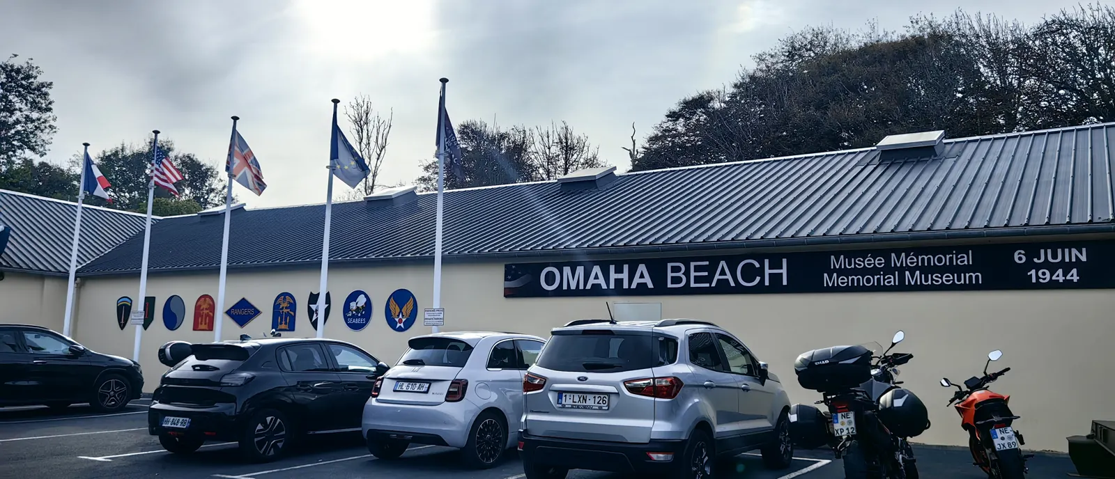 Omaha Beach Memorial Museum in Saint-Laurent-sur-Mer: façade reading Musée Mémorial 6 juin 1944 with the French, American, British and European flags and painted insignia of the 1st, 29th and 2nd Infantry Divisions, Rangers, Seabees and US Army Air Forces