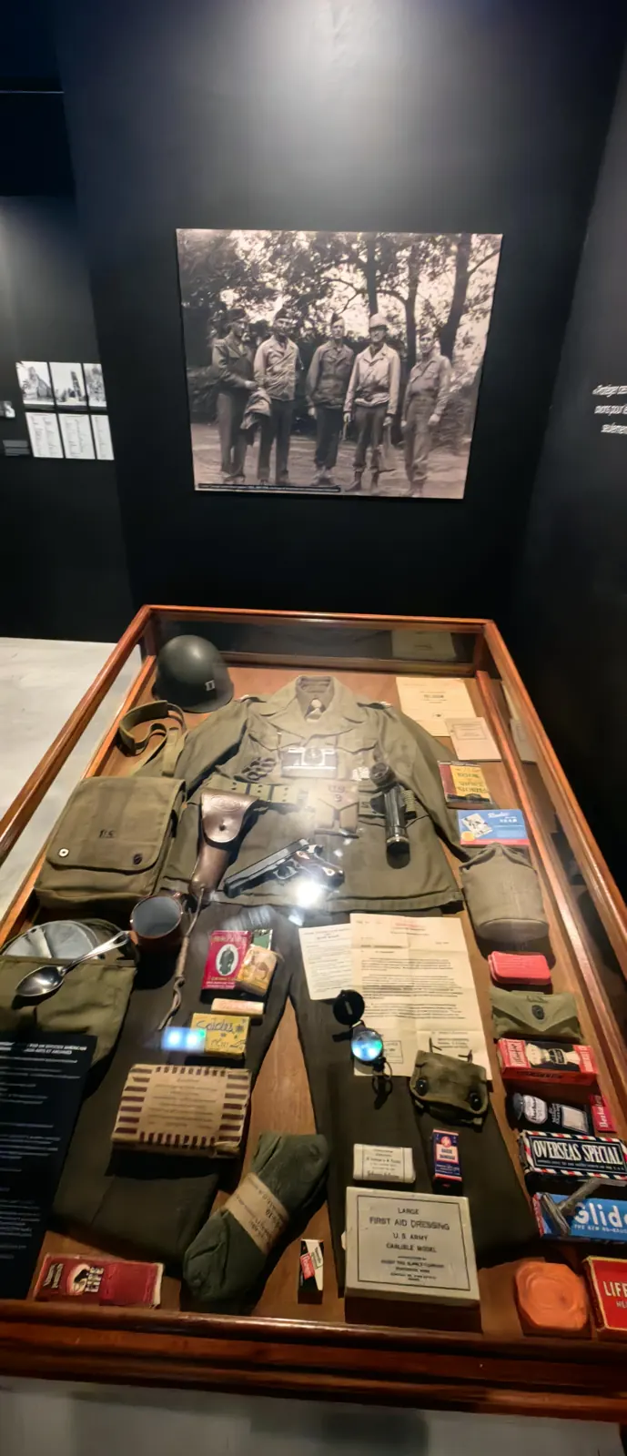 Glass case with a 1944 American infantryman's kit, helmet, field jacket, pistol and rations, under a period photograph at the Overlord Museum